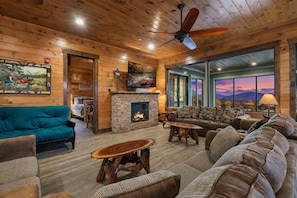 A cozy cabin interior with a stone fireplace, wooden ceiling, and a large window offering a scenic view. - Sierra Splash Lodge | Pool/Theater | Sleeps 72 (Sevierville)
