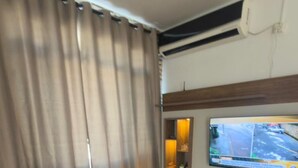A modern bedroom with a bed, curtains, a TV, and a small shelf. - Badaró's Beach Relax near the beach, 5 minutes from Mallet, shopping, and pet-friendly. (Praia Grande)