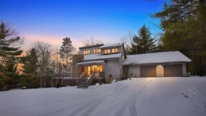 Une maison à deux étages dotée d’un porche couvert et d’un garage, entourée d’un paysage enneigé et d’arbres.