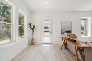 Ein geräumiger Raum mit großen Fenstern, einer hölzernen Bank, einer Tür mit Glasfeldern und einem gerahmten Kunstwerk an der Wand.