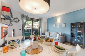 A dining area with a table set for two, a bookshelf, a sofa, and a view of the outdoors. - Blu Oltrelago: Leggiuno Lake Maggiore Apt w/ Shared Pool & Garden, Leggiuno, Italy (Leggiuno)