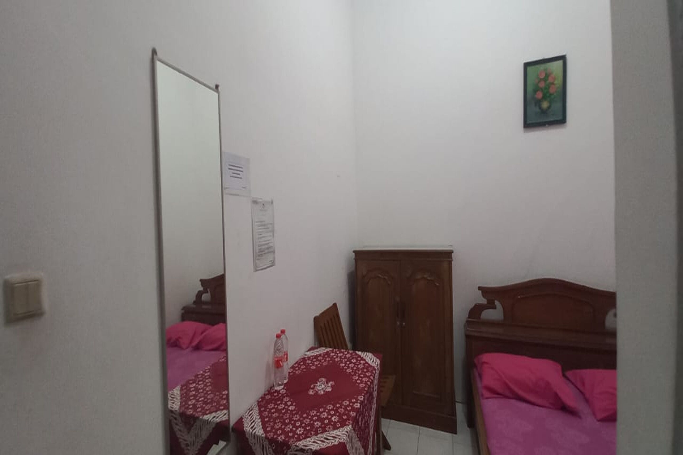 Una habitación pequeña y poco amoblada, con una cama, un armario de madera, una silla y una mesa cubierta con un mantel rojo.