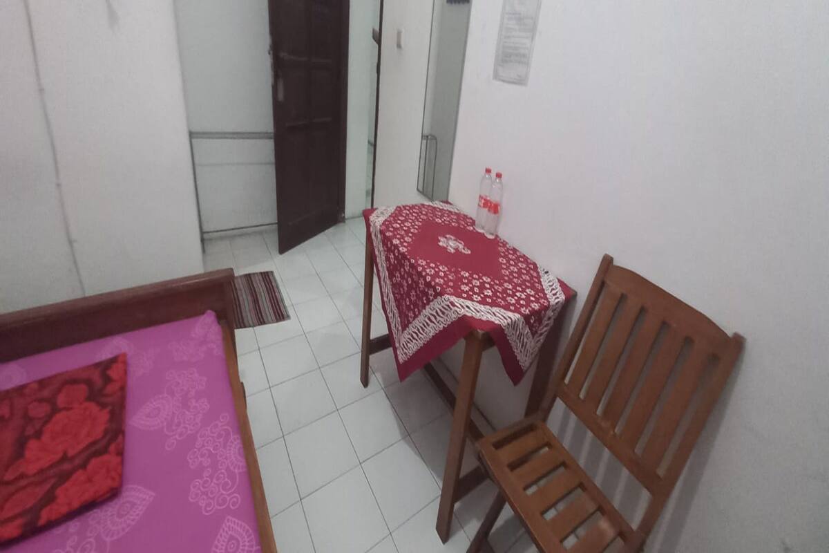 Une petite pièce avec un lit, une chaise en bois, une petite table recouverte d’une nappe rouge et une bouteille d’eau.