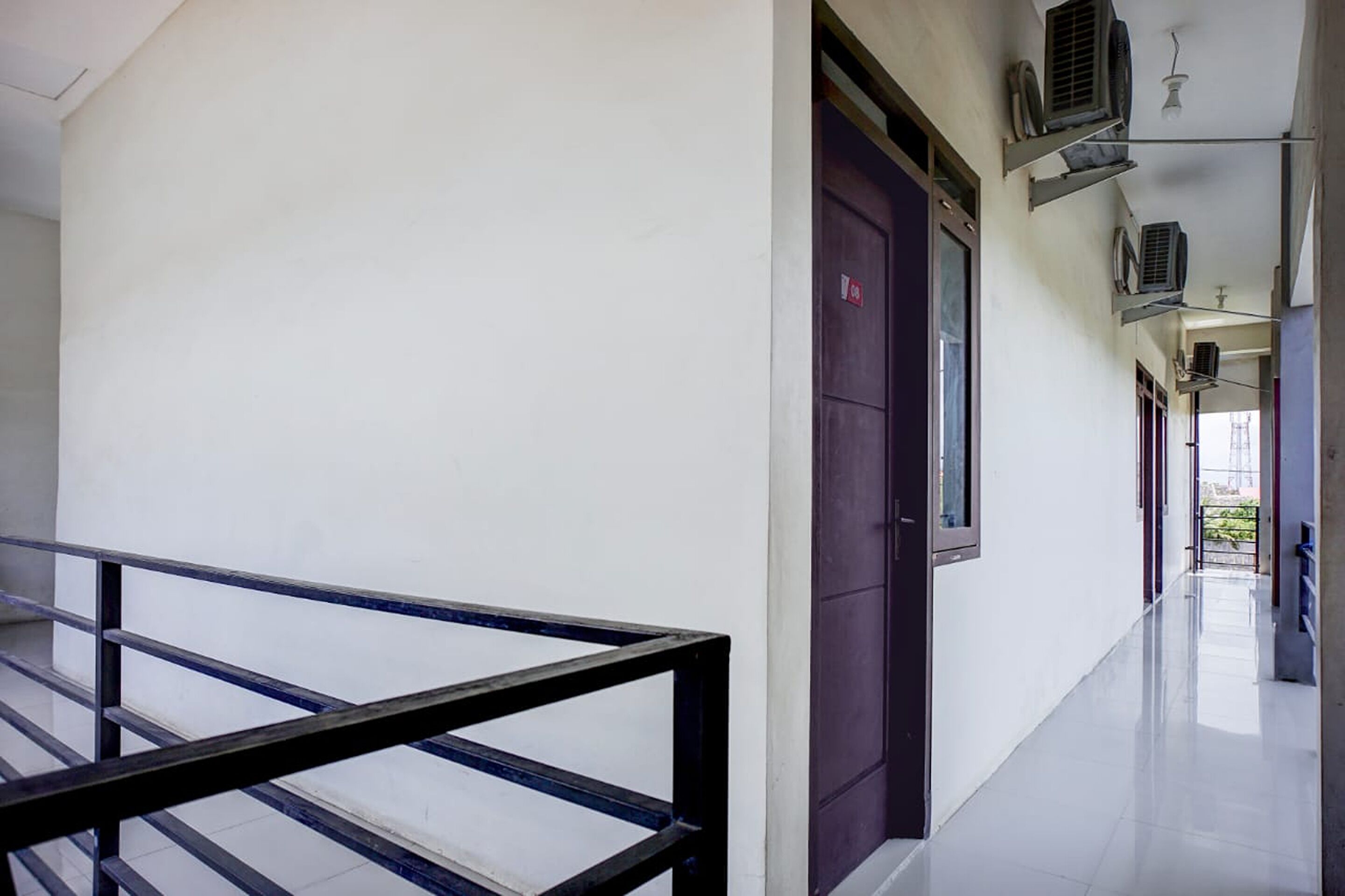 A corridor with a metal railing, a door with a red sign, and air conditioning units on the wall.