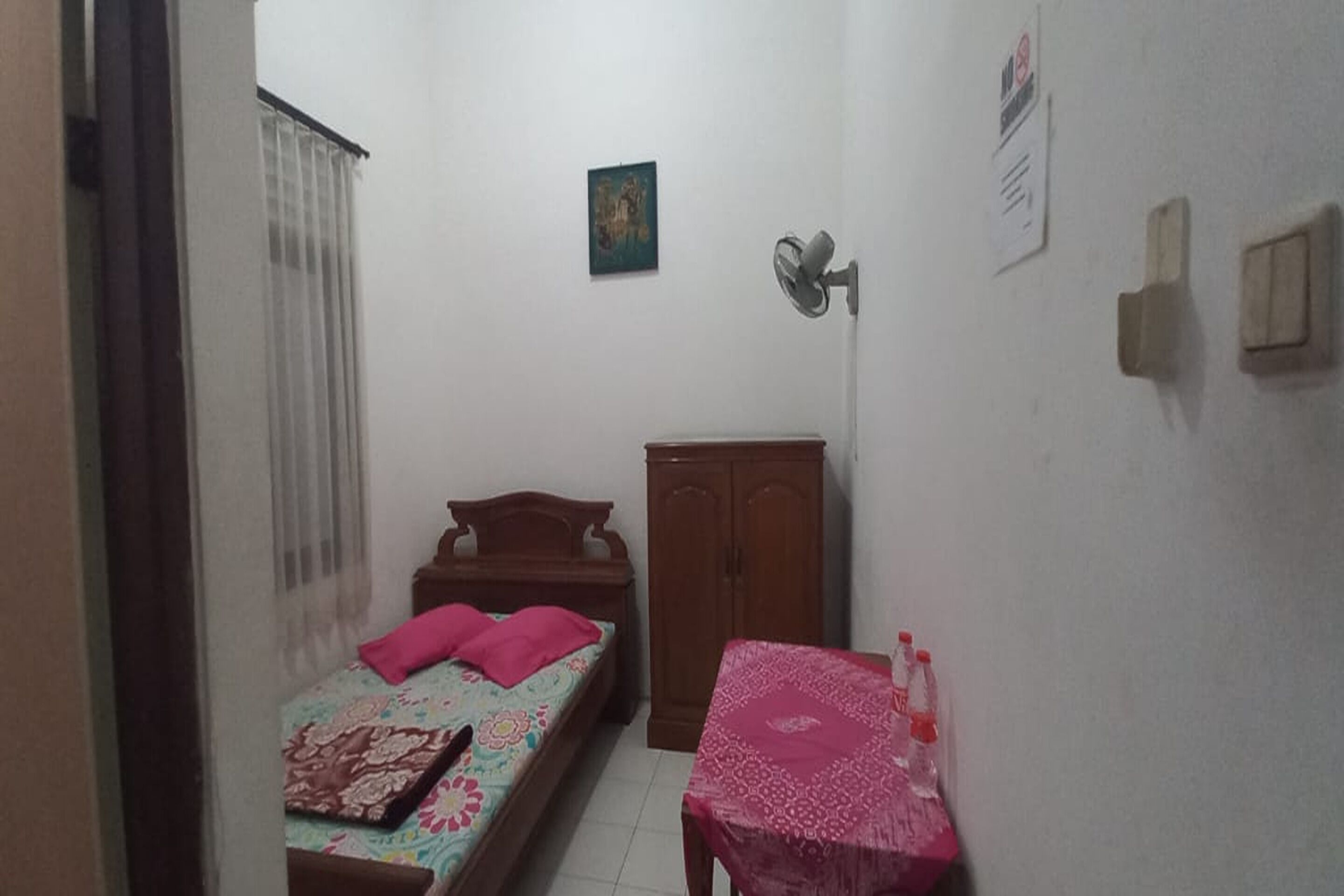 A small, dimly lit room with a single bed, a wooden wardrobe, and a table with bottles.