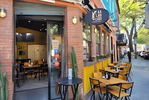An outdoor seating area with tables and chairs, a storefront with a sign, and a brick building facade. - Perto da Praia - Melhor Localização (Rio de Janeiro)