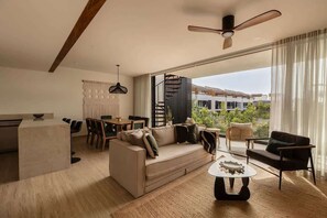 Un salon moderne avec un canapé, une table basse et des fauteuils, un coin repas avec une table et des chaises, un ventilateur de plafond et de grandes fenêtres offrant une vue sur les bâtiments et la verdure.