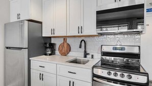 Una cocina con armarios blancos, un refrigerador de acero inoxidable, una cafetera, una tabla de cortar de madera, un fregadero y una estufa.