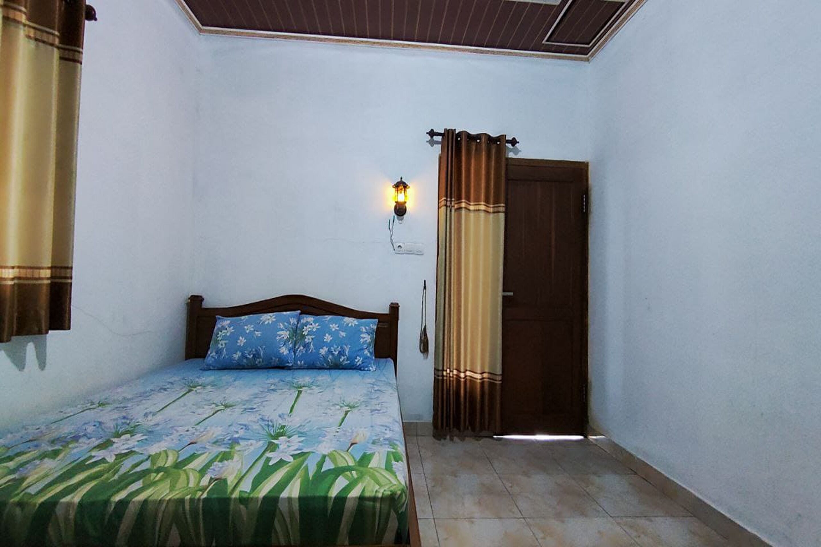 A small, well-lit bedroom with a bed, a wall-mounted light, a door with curtains, and a tiled floor.