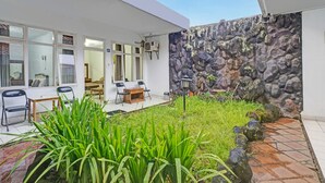A modern building with a stone wall, a patio with chairs and a table, and a garden area with plants. - OYO 95055 Homestay Panasan Pak Budi (Sambi)