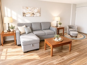 Ein Wohnzimmer mit einem grauen Ecksofa, einem Holztisch, einem grauen Sessel und zwei Nachttischen mit Lampen.