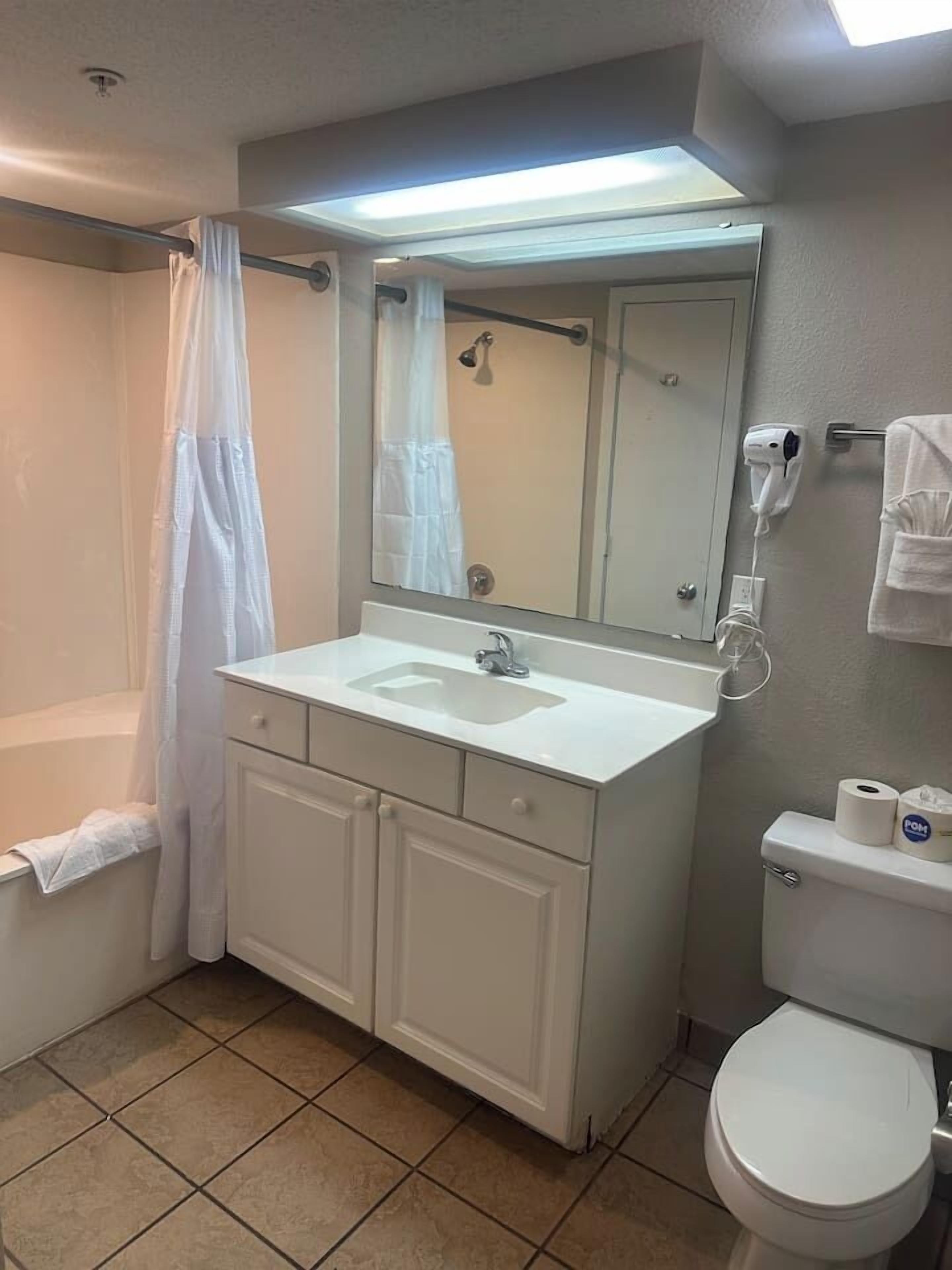 Un baño con lavamanos blanco, espejo y retrete. Hay una cortina de ducha y un perchero para toallas.