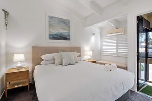 A bedroom with a large bed, two bedside tables with lamps, a ceiling fan, an air conditioning unit, and a window with blinds. - Noosa River 2BD Townhouse - Modern & Stylish (Noosa)