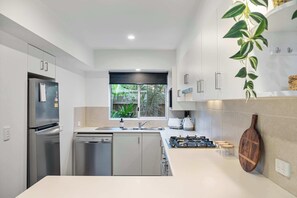 A modern kitchen with stainless steel appliances, a dishwasher, a gas stove, and a window with a view of greenery. - Noosa River 2BD Townhouse - Modern & Stylish (Noosa)