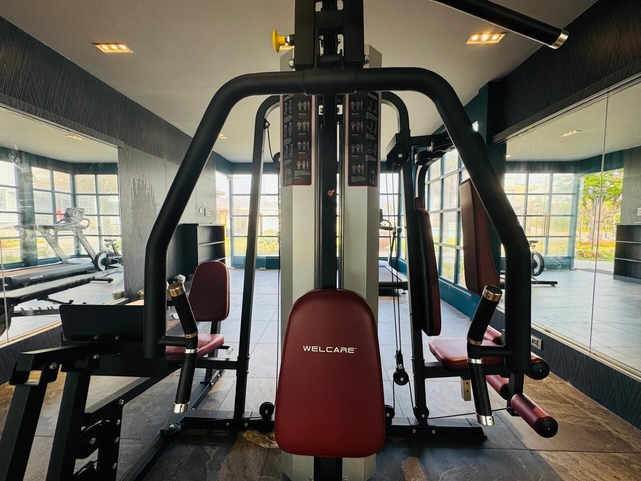 A gym with a red WELLCARE bench, weight machines, and large windows offering a view of the outdoors.
