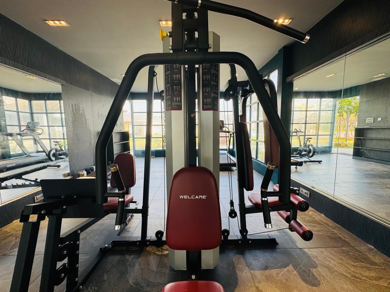 A gym with a variety of exercise machines, including a bench press and a leg press, with large windows providing natural light.