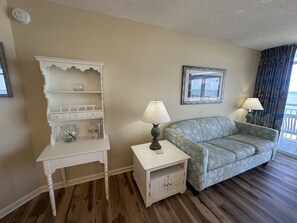 Ein Zimmer mit einem weißen Schminktisch, einem gemusterten Sofa, einem kleinen Tisch mit Lampe und einem gerahmten Bild an der Wand.