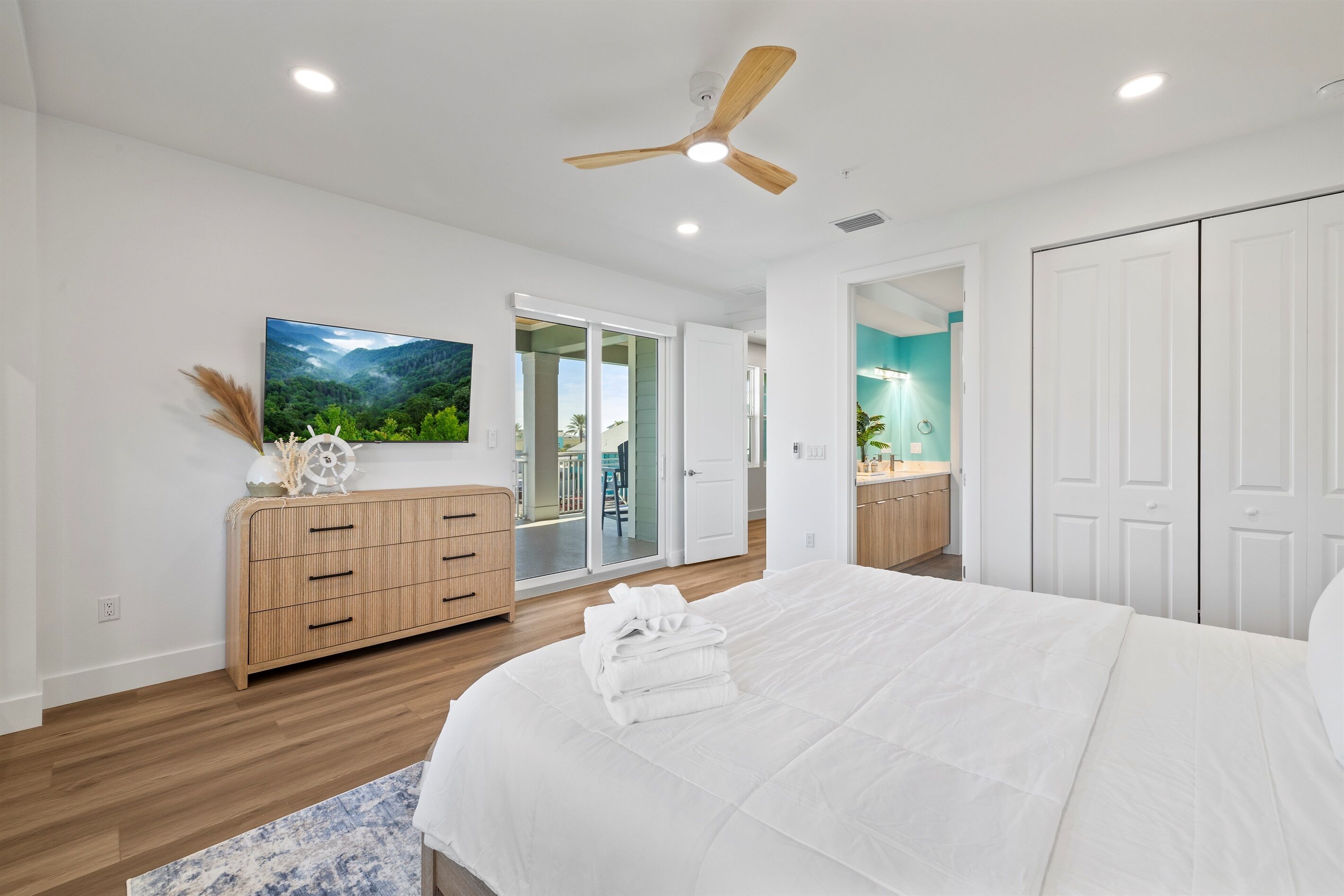 A bedroom with a large bed, a wooden dresser, a ceiling fan, and a view of the outside.