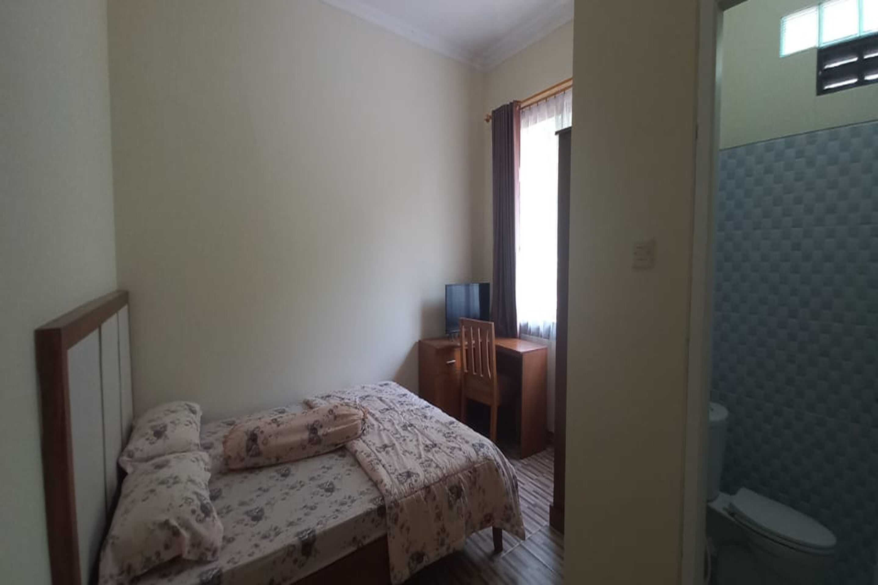 A bedroom with a bed, a desk, and a chair. There is a window with curtains and a view of a bathroom with a toilet.