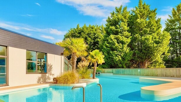 A modern pool area with clear blue water, surrounded by lush greenery and a contemporary building. - Private House in the Normandy Garden – Pierre & Vacances Resort (Branville)