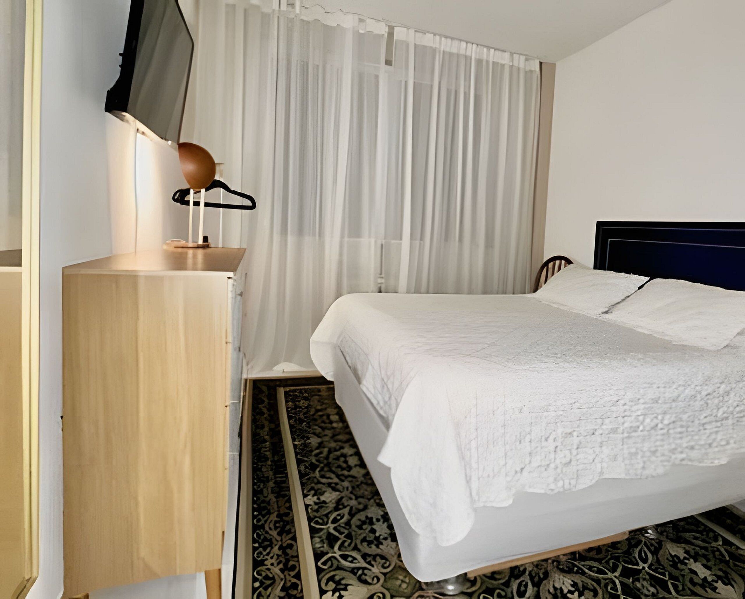 Een hotelkamer met een bed, een houten dressoir, een televisie op een standaard en een raam met dunne gordijnen.