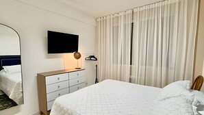 Une chambre d’hôtel avec un lit, une commode, un téléviseur fixé au mur et une fenêtre avec des rideaux légers.