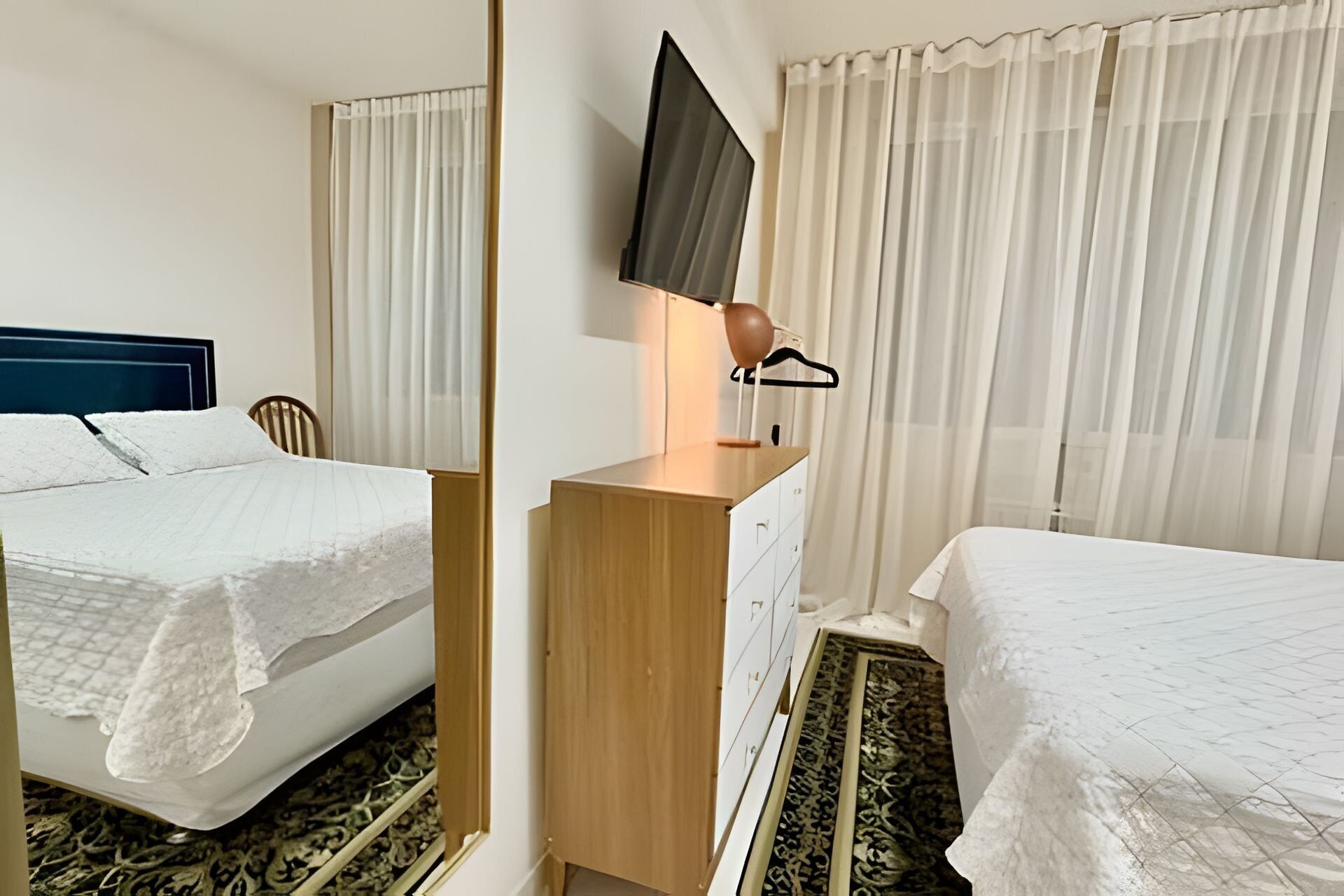 A hotel room with two beds, a TV mounted on the wall, a wooden dresser, and a window with sheer curtains.