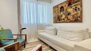 Un salon avec un canapé blanc, des fauteuils bleus, un tapis à rayures et un grand tableau représentant une scène historique.