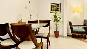 Un coin repas avec une table blanche et quatre chaises en bois dotées de coussins blancs. Une plante en pot et une lampe se trouvent sur le côté droit.