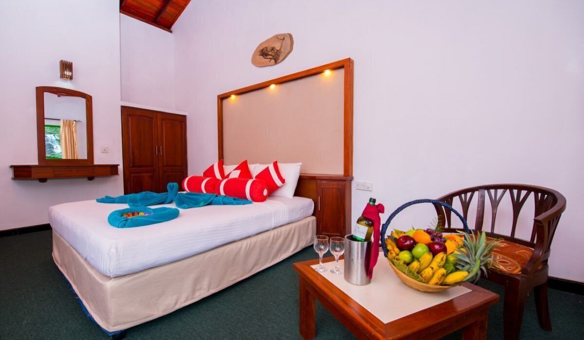 A hotel room with a bed, a wooden chair, a table with a fruit basket, and a bottle of champagne.
