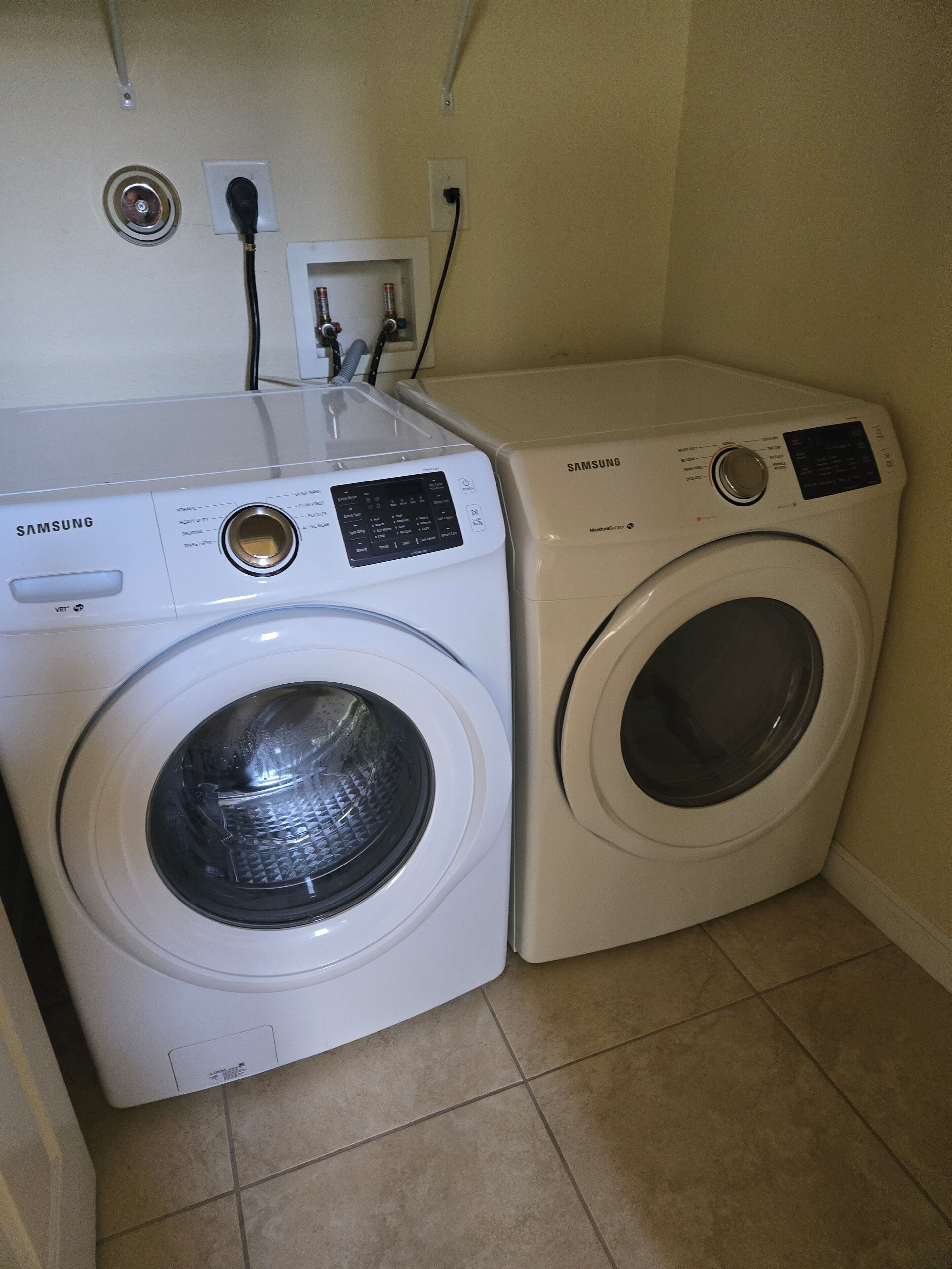 Two white Samsung washing machines in a corner with tiled flooring.