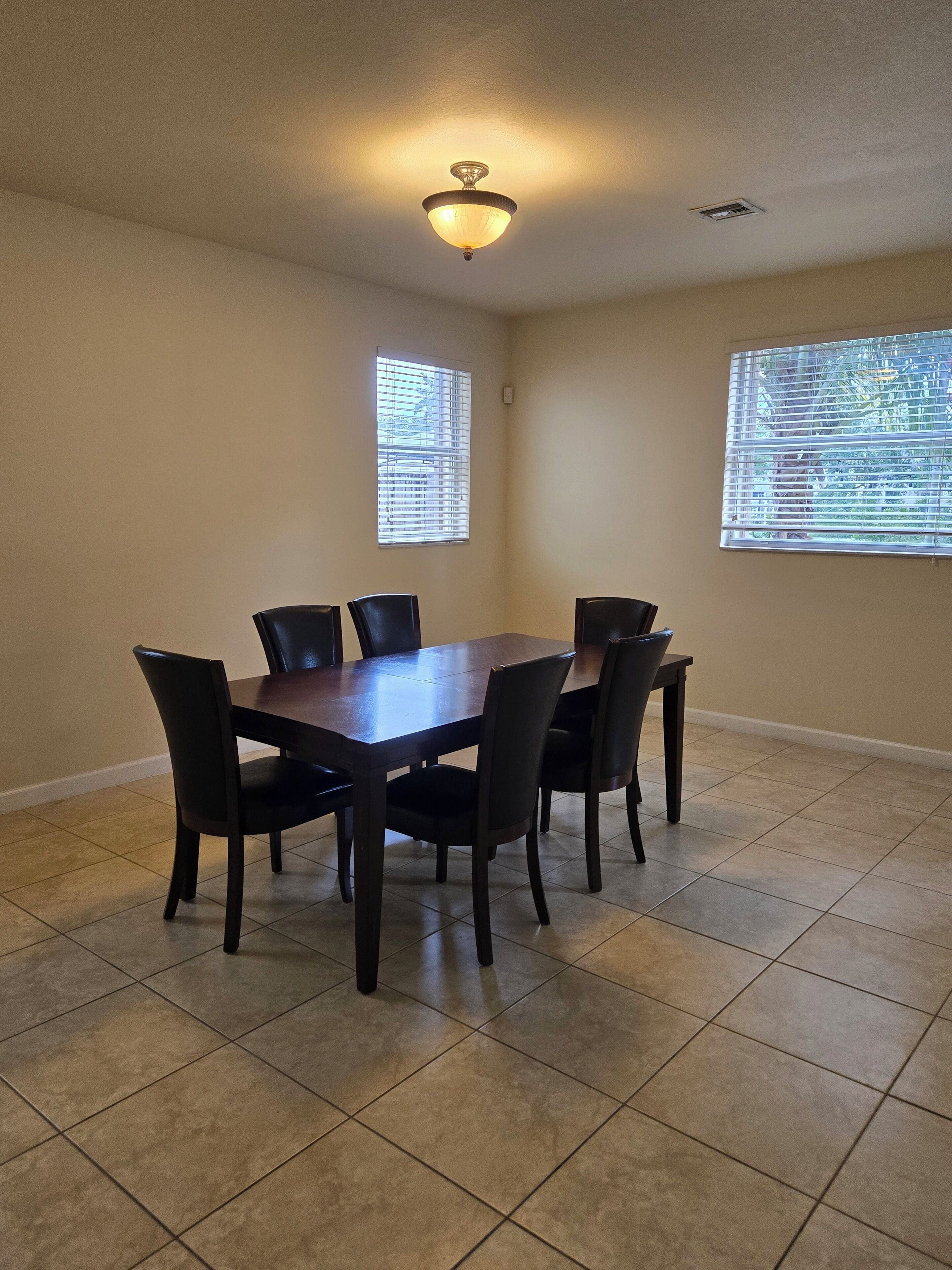 Une salle à manger avec une table rectangulaire et six chaises, deux fenêtres équipées de stores et un luminaire au plafond.