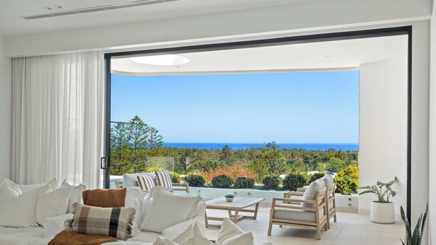 Una sala de estar moderna con sofá, sillones y una mesa de centro, que da a un balcón con vistas al mar y al follaje.