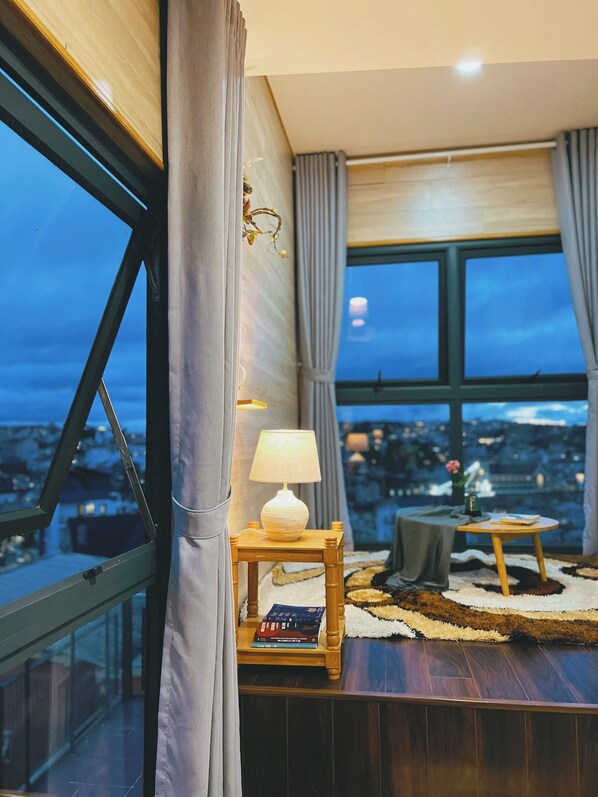 A modern living room with a view of a cityscape through large windows, a small wooden side table with a lamp, and a cozy rug.