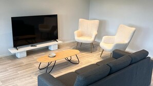Ein modernes Wohnzimmer mit einem grauen Sofa, zwei weißen Sesseln, einem Holztisch und einem Flachbildfernseher auf einem weißen Schrank.