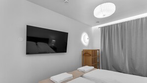 Une chambre d’hôtel moderne dotée d’un grand téléviseur à écran plat fixé au mur, d’un lit avec des serviettes blanches soigneusement pliées, d’une armoire en bois et d’une grande fenêtre avec des rideaux gris.