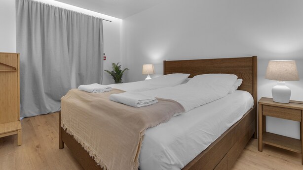 Un lit double bien fait, avec une couverture beige, une table de chevet avec une lampe et un rideau gris.