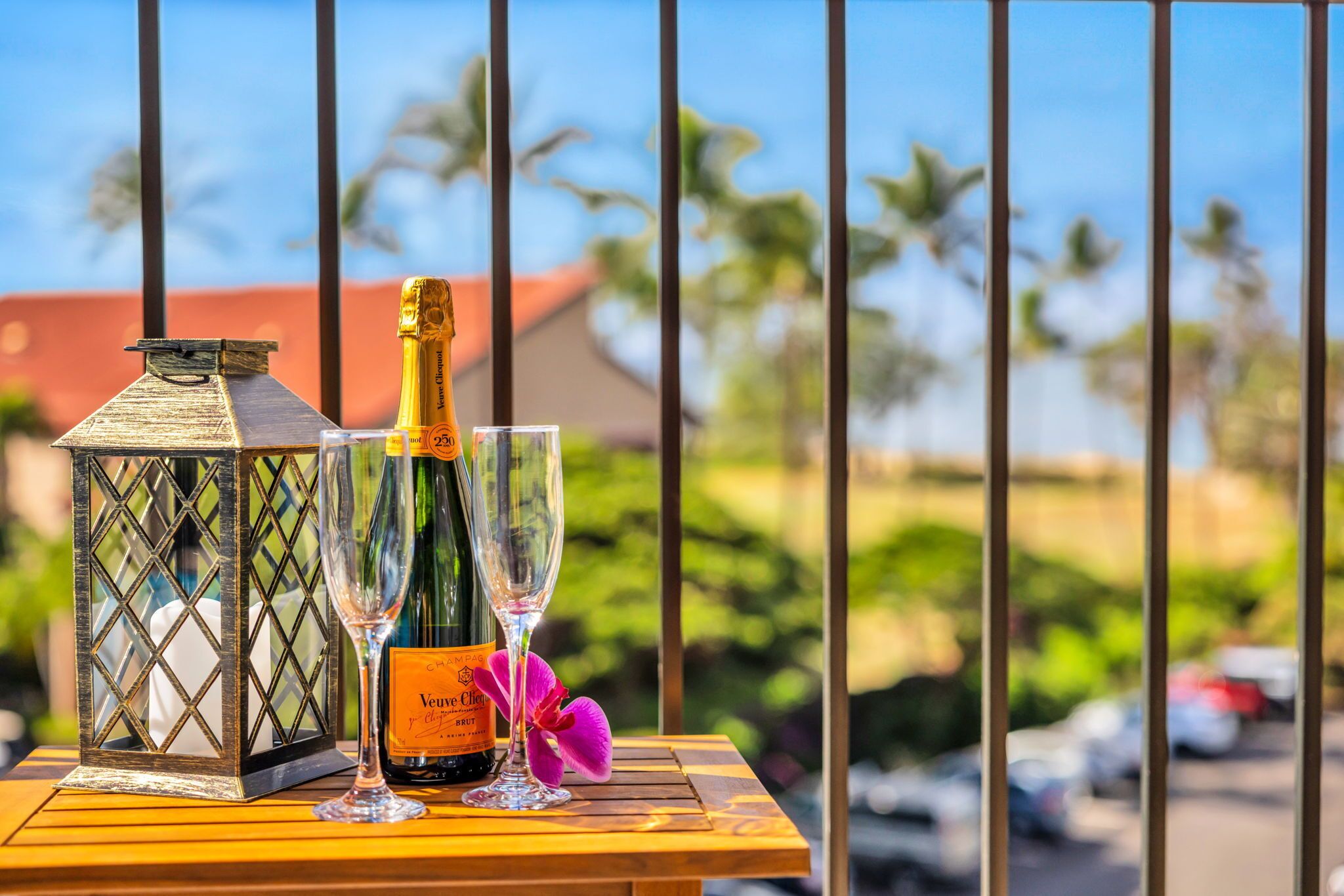 A bottle of Veuve Clicquot champagne, a glass, and a lantern on a wooden table with a view of palm trees and a building in the background.