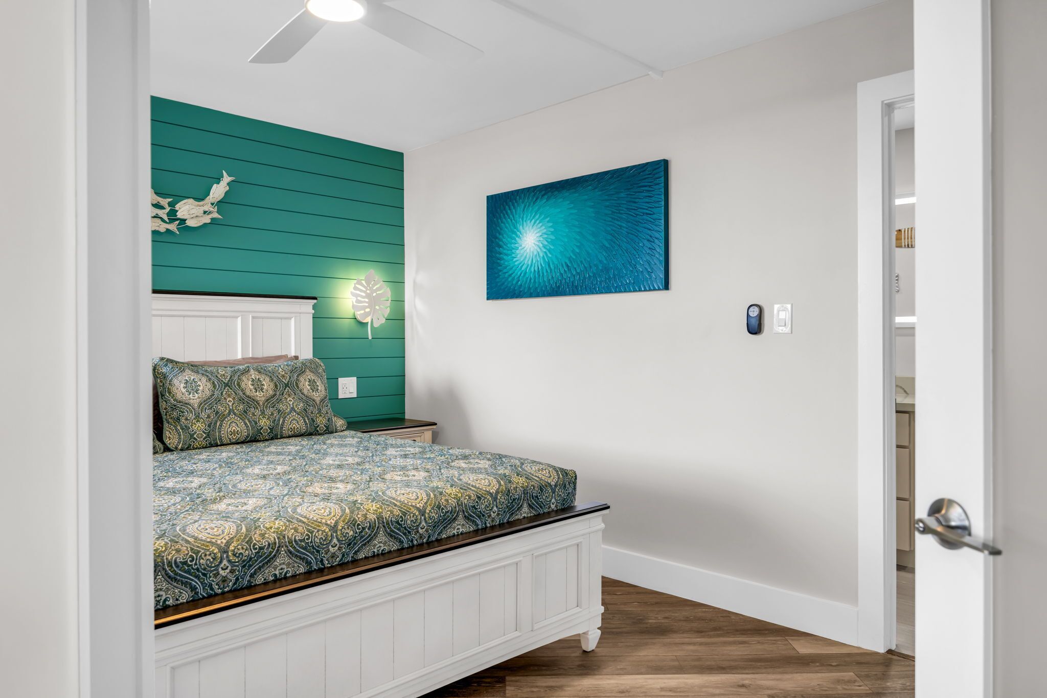 A bedroom with a bed, a ceiling fan, a wall-mounted light, and a painting.