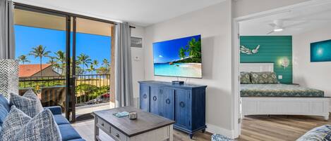 Una sala de estar con un televisor grande, un sofá azul, una mesa de centro y vistas a palmeras y a la playa a través de puertas correderas de vidrio.