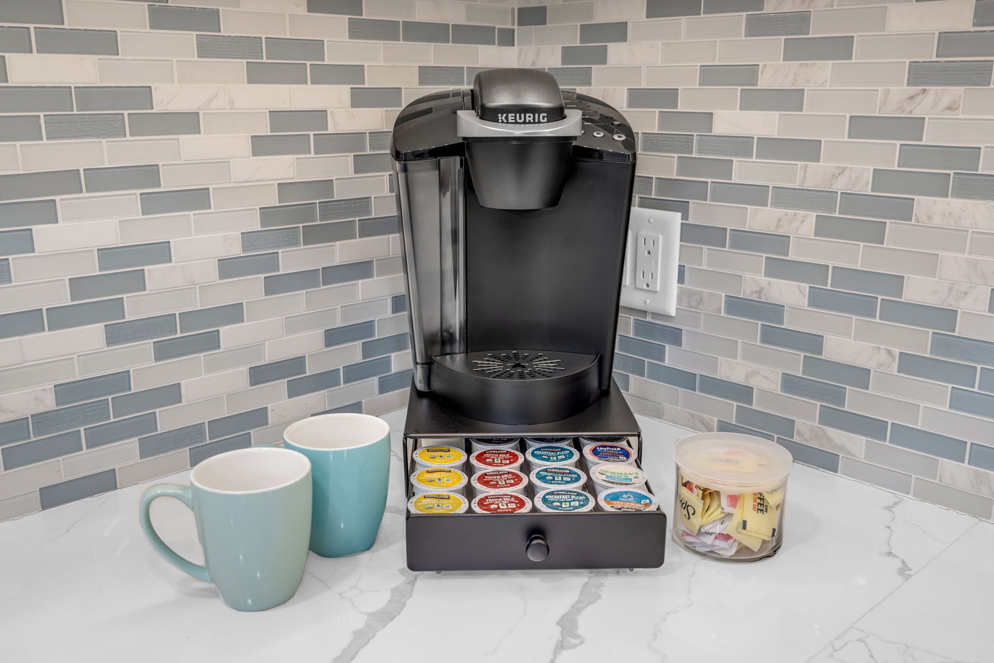 Eine Kaffeemaschine von Keurig mit Pod-Halter, zwei blaue Tassen und einer Kaffeepad-Verpackung auf einer Küchenarbeitsplatte.