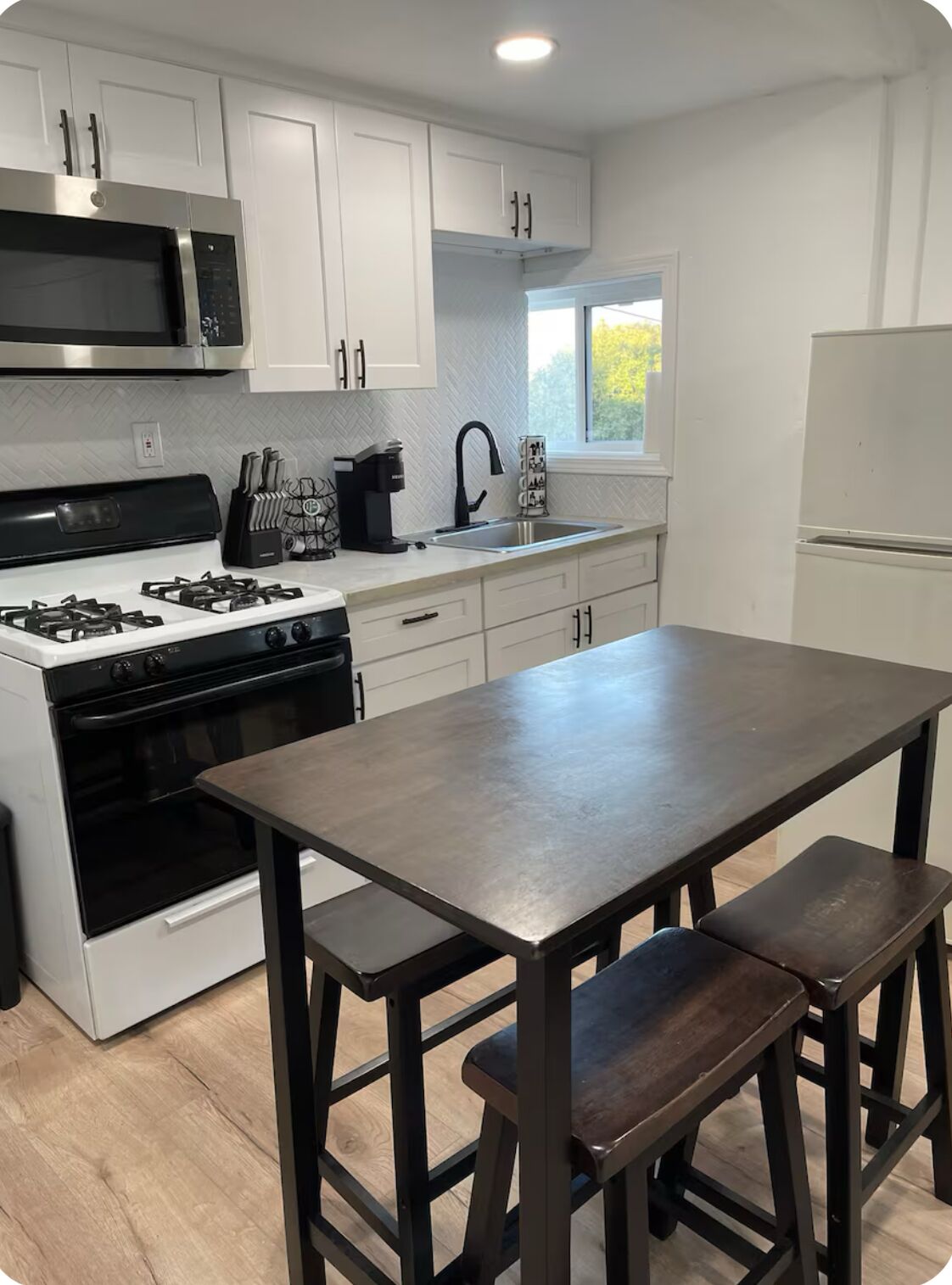 A modern kitchen with white cabinets, a stainless steel stove, a microwave, and a refrigerator. There is a window providing natural light.