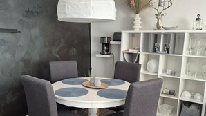 Et spiseområde med et rundt bord, fire grå stole, en hvid lysekrone og et hvidt reolsystem med glas og dekorative genstande.