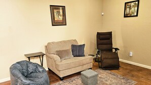 Un salon avec un canapé beige, un fauteuil inclinable marron, un pouf gris et un tapis à motifs.