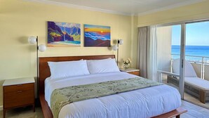 Een ruime slaapkamer met een groot bed, twee schilderijen, een nachtkastje en een bijzettafeltje, en uitzicht op de oceaan door een glazen schuifdeur.