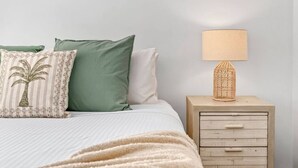 Una cama con almohadas verde y blancas, un plaid color beige y una mesita de noche de madera con una lámpara tejida.