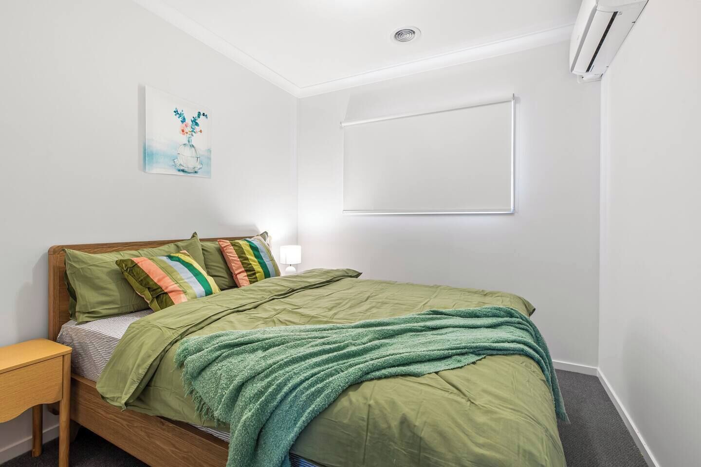 A bedroom with a bed, pillows, a bedside table, a lamp, a wall-mounted air conditioner, and a painting.