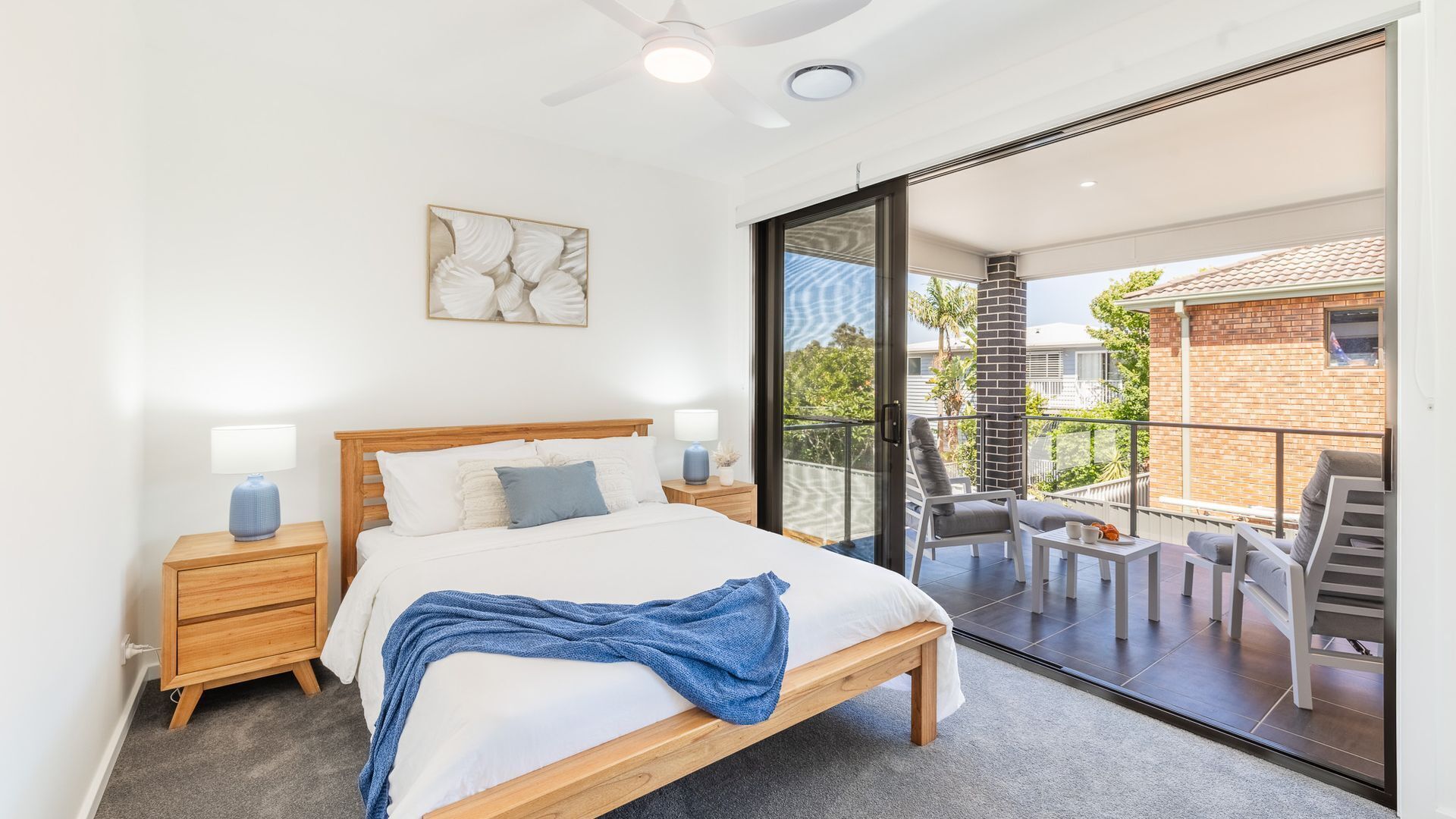 A bedroom with a bed, bedside tables, a ceiling fan, and a large sliding glass door leading to an outdoor balcony with a chair and table.