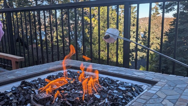 Ein Feuerkorb mit brennenden Holzscheiten, umgeben von Steinplatten, auf einem Balkon mit Geländer und Bäumen im Hintergrund.