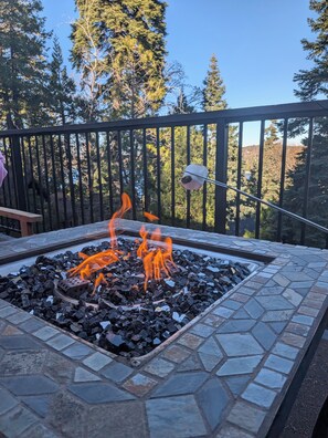 A fire pit with burning logs surrounded by stone tiles, set on a balcony with a railing and trees in the background. - Beautiful 3-bedroom house in tranquil Lake Arrowhead getaway (Lake Arrowhead)